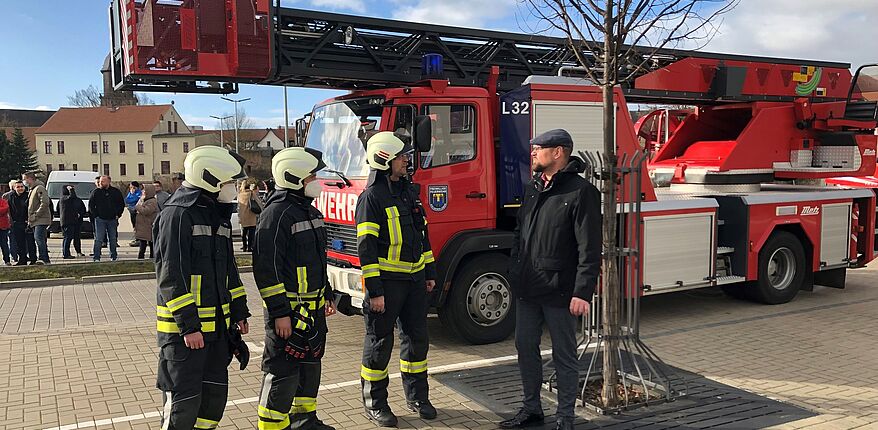 Auf dem Foto sind Kameraden der Freiwilligen Feuerwehr vor eine Einsatzfahrzeug im Gespräch mit Landrat Martin Stichnoth zu sehen. Den Hintergrund des Fotos bietet einen Blick in Richtung Stadtzentrum von Haldensleben.