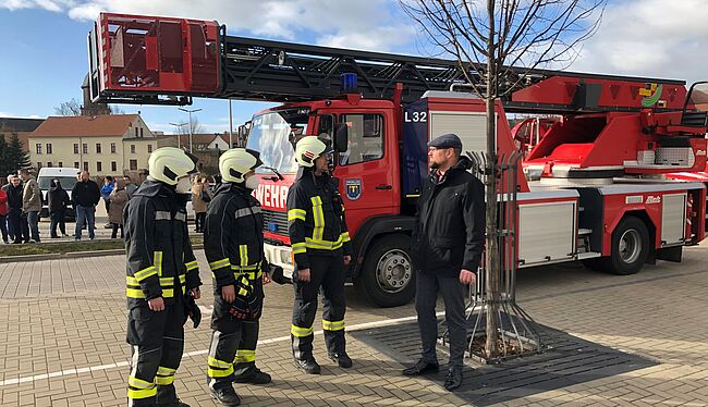 Auf dem Foto sind Kameraden der Freiwilligen Feuerwehr vor eine Einsatzfahrzeug im Gespräch mit Landrat Martin Stichnoth zu sehen. Den Hintergrund des Fotos bietet einen Blick in Richtung Stadtzentrum von Haldensleben.