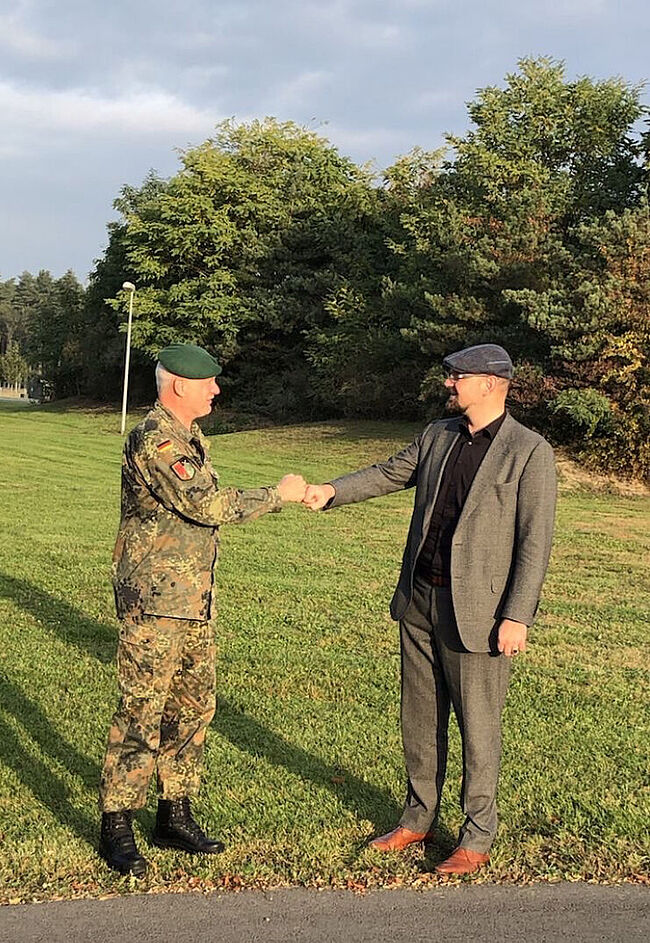 Das Foto zeigt Landrat Martin Stichnoth und Oberst Michael Knoke bei der Gratualtion in Letzlingen. Beide Herren begrüßen sich mit der "Faust". Sie stehen auf einer Wiese. Im Hintergrund ist ein Wald zu sehen. 